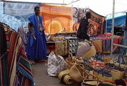 A vendor in a blue robe beside a stall of rugs, fabrics and baskets at Waterlooplein